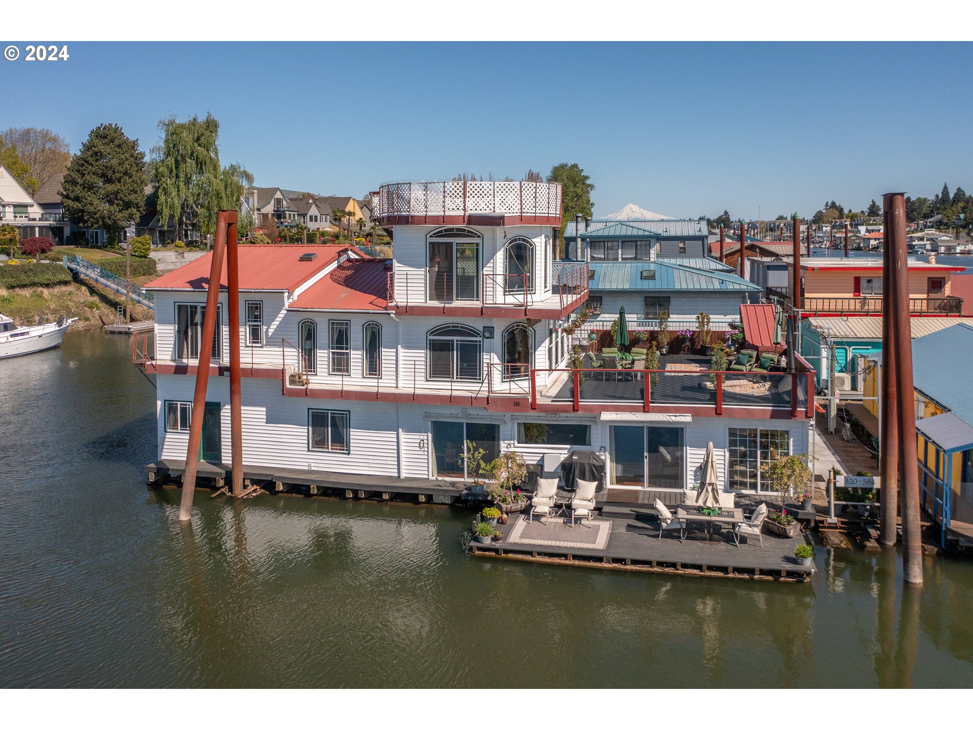 A truly exceptional floating home located in Tomahawk Moorage, nestled in a prime west end slip offering breathtaking sunset views from decks spanning three floors. This residence features two fully equipped living areas; the current owner enjoys both, though one could easily serve as a separate guest or rental space. Upon entering through an enclosed foyer, you’re welcomed into a spacious entertaining area complete with a gourmet kitchen showcasing a freestanding luxury gas range, pot filler, granite countertops, an extended eating bar, and an impressive wall of cabinetry. West-facing windows perfectly frame the river and sunset vistas. This level also includes a gas fireplace, generous living and dining spaces, a large bedroom, and a full bathroom. Ascend the wooden spiral staircase to the second floor to find a cozy family room with a gas stove, panoramic river views, a sizable private deck, and a second full kitchen equipped with a full-size fridge, stove, and built-in dishwasher. The primary bedroom on this floor boasts vaulted wood-paneled ceilings, a walk-in closet, and a jetted bathtub, creating a serene private retreat. An additional non-conforming bedroom is also located here. The third floor is accessible via another spiral staircase leading to a spacious cupola room, ideal for meditation, yoga, or quiet reflection, featuring stunning views of Mt. Hood and its own private deck. This unique and spacious floating home offers ample room and private areas for family and friends to enjoy resort-style river living. The boat well accommodates large vessels, perfect for skiing or fishing. Combining traditional charm with modern amenities, this home truly stands out.